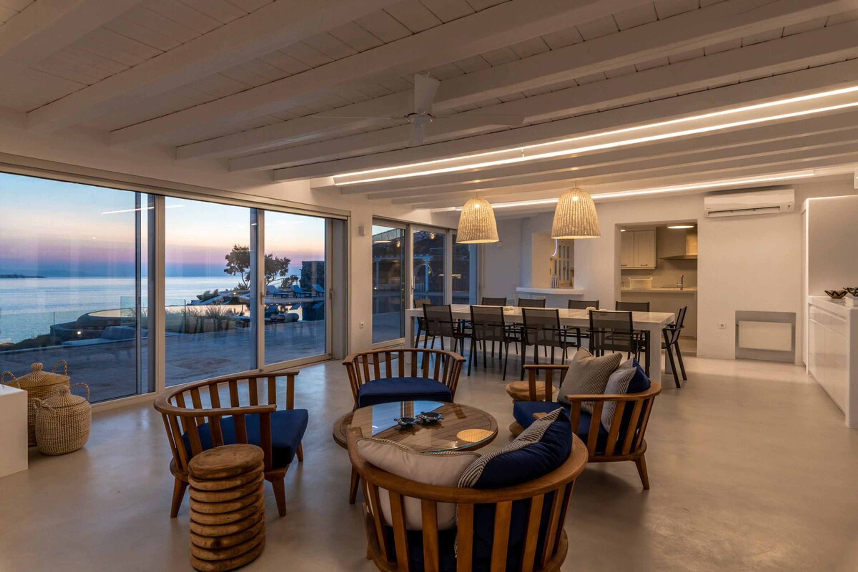 A modern interior with a dining area, seating with cushions, and large windows overlooking a body of water.
