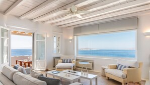 A modern living room with a large window overlooking the sea, a ceiling fan, and a cozy seating area. - Luxury Villa Roshell (Mykonos)