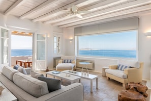 Ein modernes Wohnzimmer mit großem Fenster zum Meer, einem Deckenventilator und einer gemütlichen Sitzecke.