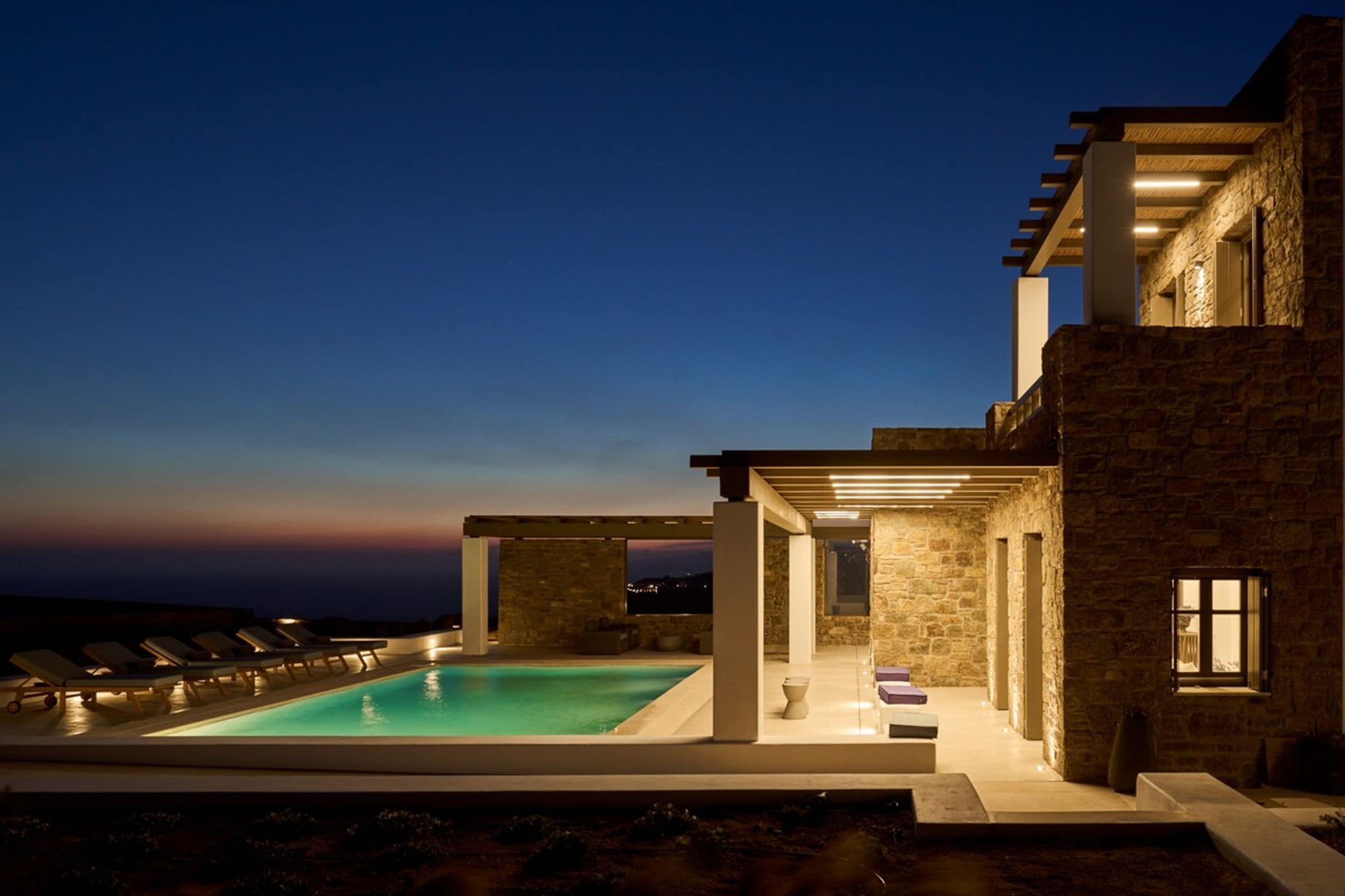 Un espace moderne aménagé au bord de la piscine, avec des chaises longues, un bâtiment en pierre et une vue dégagée sur le ciel.