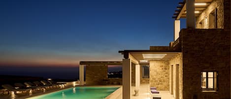Un espace moderne aménagé au bord de la piscine, avec des chaises longues, un bâtiment en pierre et une vue dégagée sur le ciel.