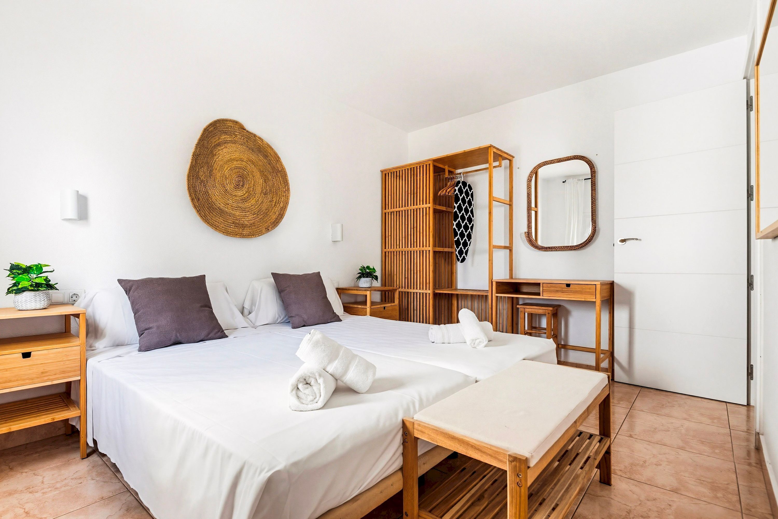A hotel room with a bed, a wooden wardrobe, a desk, a mirror, and a woven wall hanging.