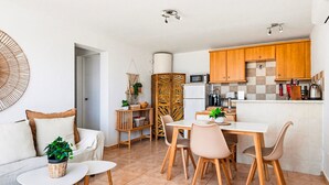 Een compacte keuken met houten kastjes, een magnetron en een koffieapparaat. Er is een eethoek met een tafel en stoelen, en een klein zithoekje met een bank en een salontafel.