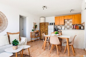 Eine kompakte Küche mit Holzschränken, einer Mikrowelle und einer Kaffeemaschine. Ein Essbereich mit Tisch und Stühlen lädt zum Verweilen ein, daneben befindet sich eine kleine Sitzecke mit Sofa und Couchtisch.