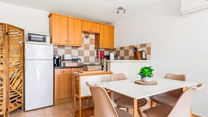 Een kleine keuken met houten kastjes, een magnetron, een koelkast, een eettafel met stoelen en een potplant.