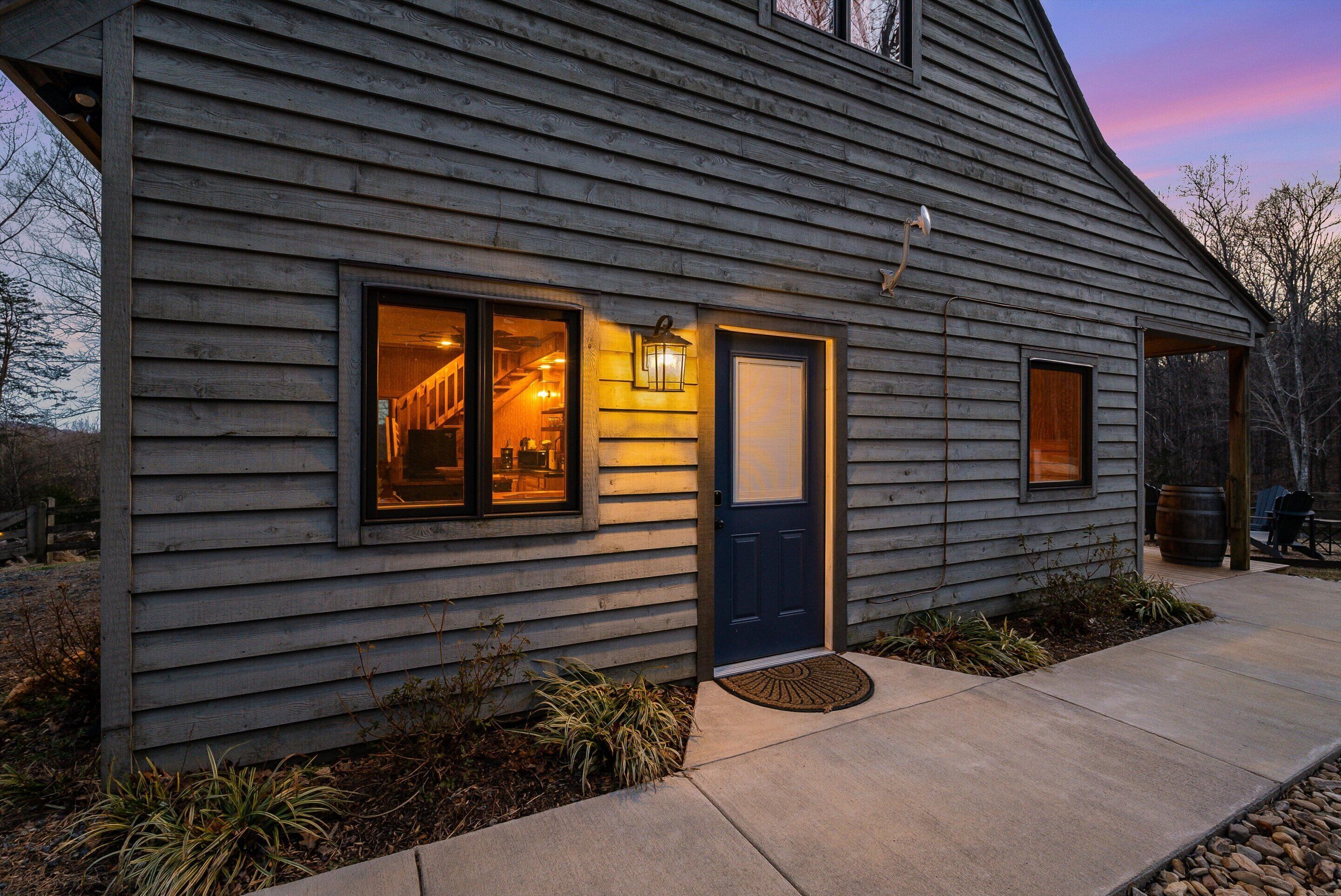 Une maison en bois avec une porte bleu foncé, un intérieur éclairé visible à travers une fenêtre et un chemin en béton menant à l’entrée.