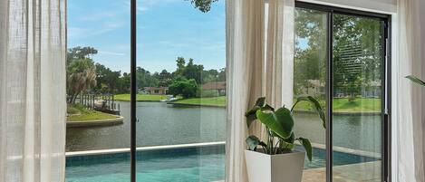 Una habitación con un amplio ventanal corredera que da a una piscina, una planta en maceta y vistas a un lago con un campo de golf.