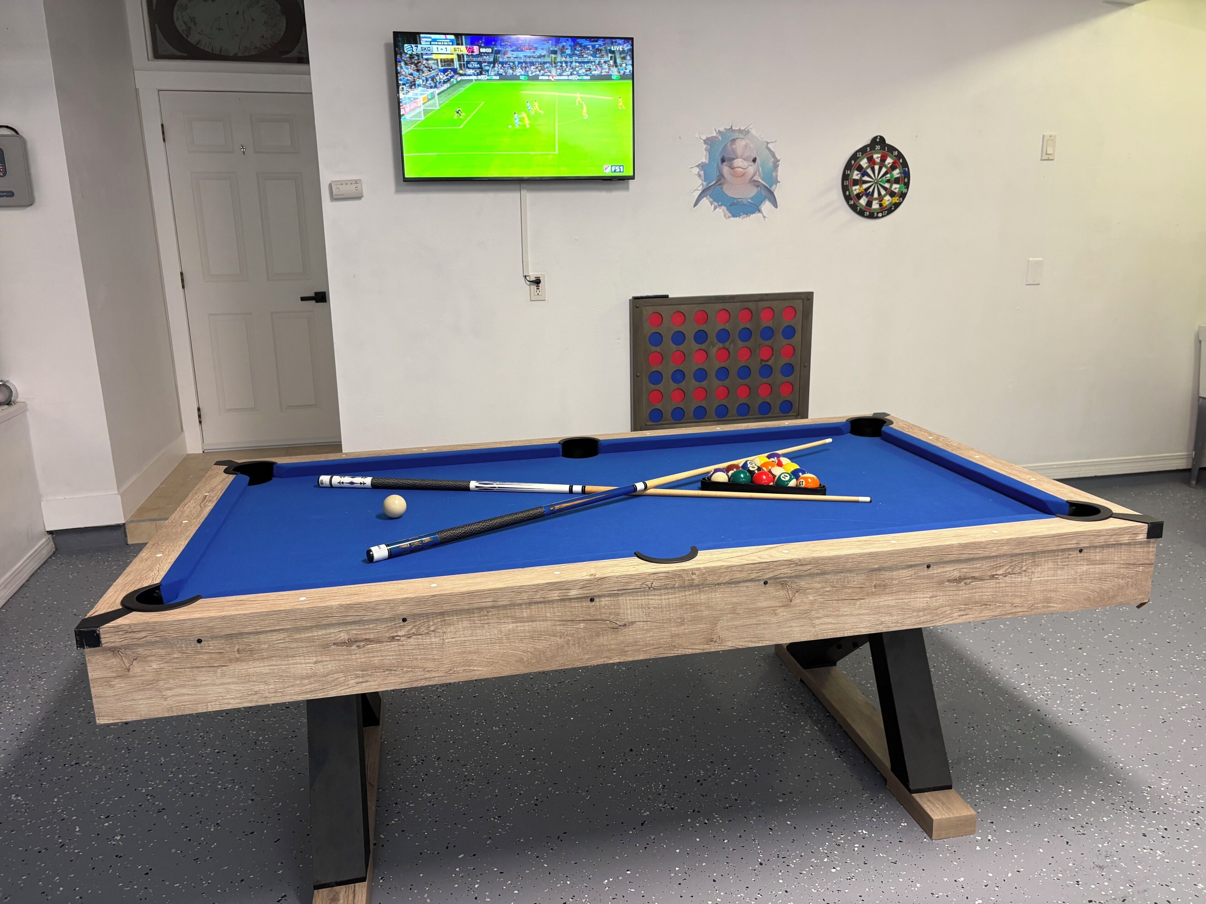 A pool table with balls and cues, a dartboard, and a television displaying a sports game.