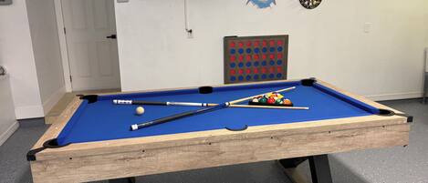 A pool table with balls and cues, a dartboard, and a television displaying a sports game.