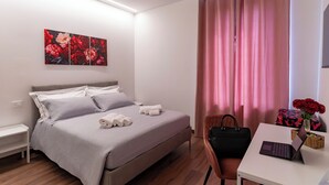 Habitación de hotel con cama, escritorio con una laptop, silla, florero con flores y una obra de arte en la pared.