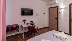 Habitación de hotel con una cama, un escritorio con una computadora portátil, un televisor mostrando 'NETFLIX', una silla roja, una maleta y una puerta.