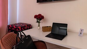Una habitación con un escritorio blanco, una laptop, un bolso negro, un televisor que muestra 'NETFLIX', un florero con flores rojas y un letrero con un código QR.
