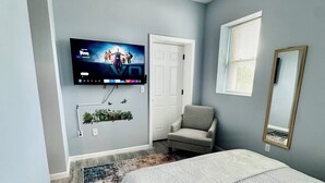 Un dormitorio con cama, televisor montado en la pared, una silla, una puerta y una ventana con persianas.