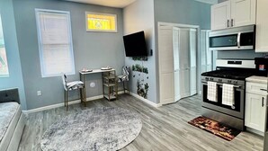 Una cocina moderna con armarios blancos, una estufa negra y un microondas. Hay una zona de comedor con una mesa y sillas, y un ventanal con persianas.