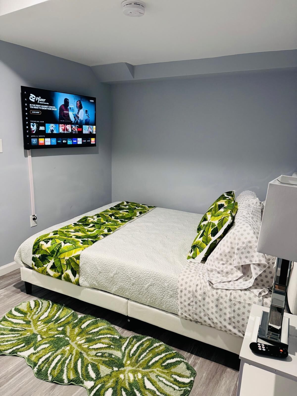 Une chambre à coucher avec un lit, un téléviseur fixé au mur, une table de chevet avec une lampe et un tapis à motifs.