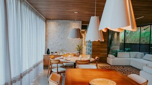 Un espace de salle à manger moderne avec une table en bois, des chaises blanches, des luminaires suspendus et un canapé en arrière-plan.
