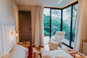 A bedroom with two beds, a chair, a table, and a large window with a view of greenery.