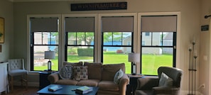 Ein Wohnzimmer mit einer Couch, zwei Sesseln, einem Couchtisch und einer Lampe. Große Fenster bieten Blick auf eine Rasenfläche und ein Haus.