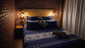 Een kamer met een bakstenen muur, een houten hoofdbord, twee bedlampjes, een bed met een decoratieve spreideken en een wit gordijn.