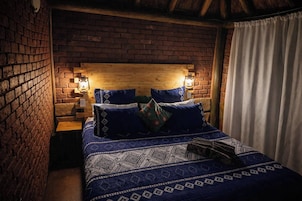 Una habitación con pared de ladrillo, cabecera de madera, dos lámparas de noche, colcha estampada y cortina blanca.