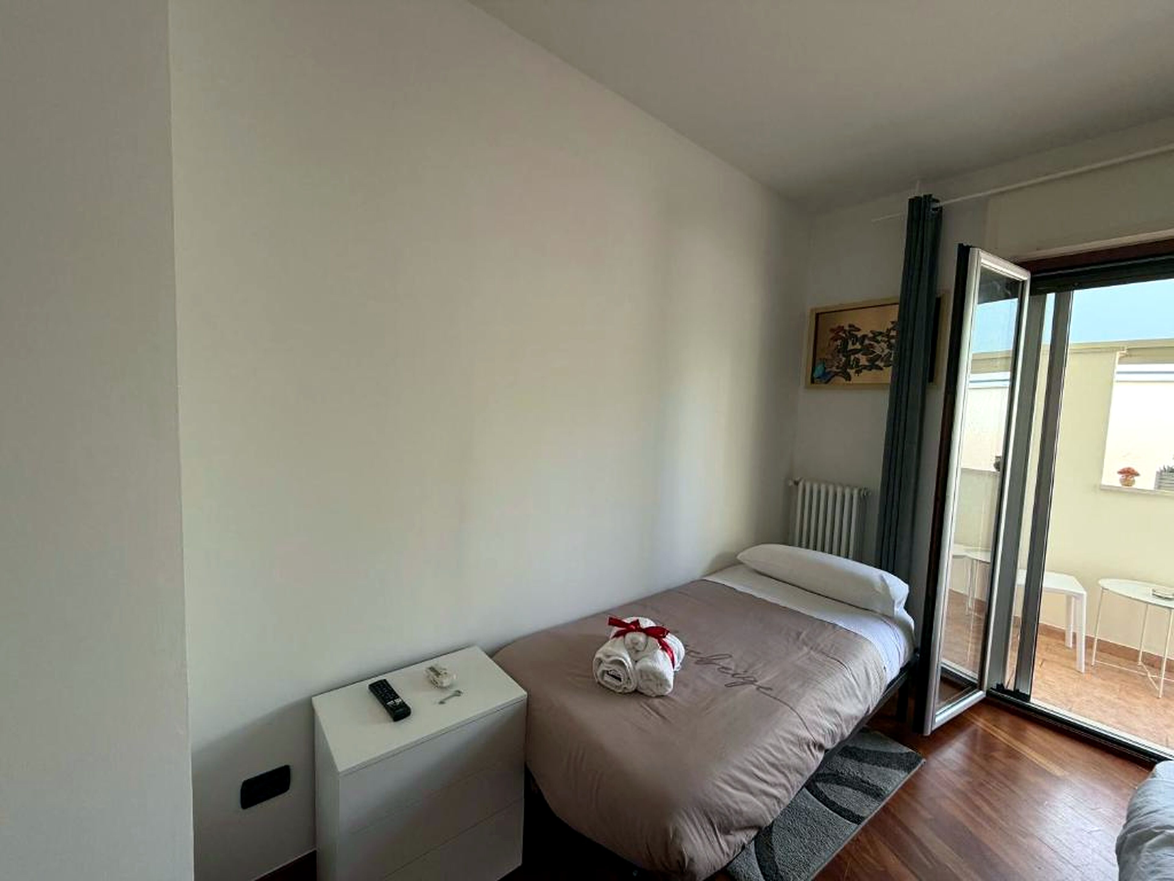 Ein Einzelbett mit weißem Kopfteil, ein Nachttisch mit Fernbedienung und ein Fenster mit Blick auf einen Balkon.