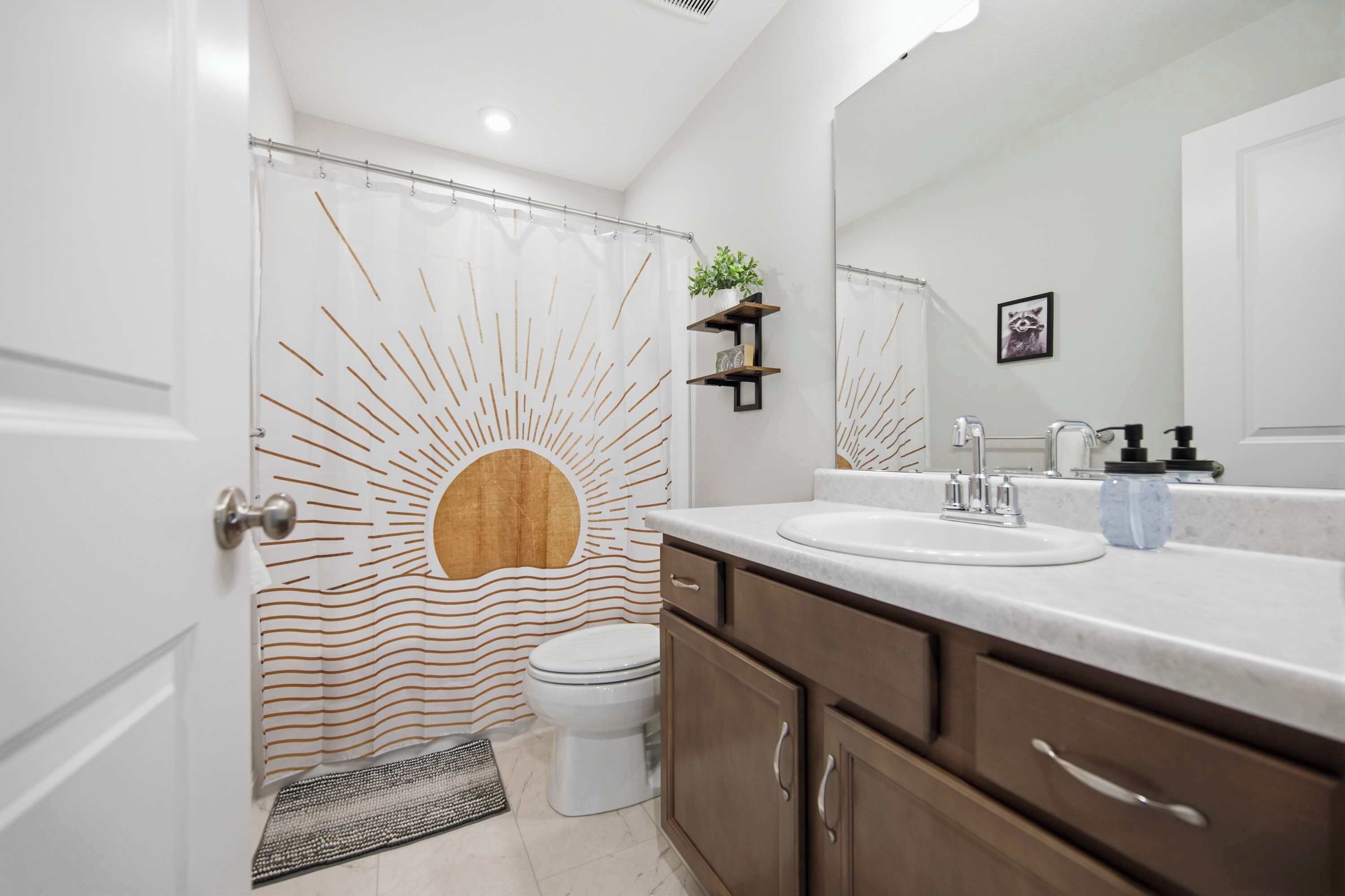 A modern bathroom with a shower curtain featuring a sunburst design, a toilet, a sink with a mirror, and a small shelf with a plant.