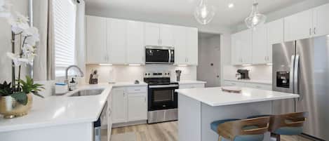 A modern kitchen with white cabinetry, stainless steel appliances, a central island with bar stools, and pendant lighting.