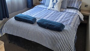 Een net opgemaakt bed met gestreept beddengoed en twee blauwe handdoeken, een nachtkastje met een lamp, en een raam met gordijnen.