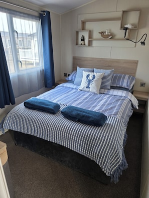 A neatly made bed with striped bedding and two blue towels, a bedside table with a lamp, and a window with curtains.