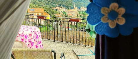 A balcony with a wicker chair, a table with a floral tablecloth, and a view of a town with buildings and hills.
