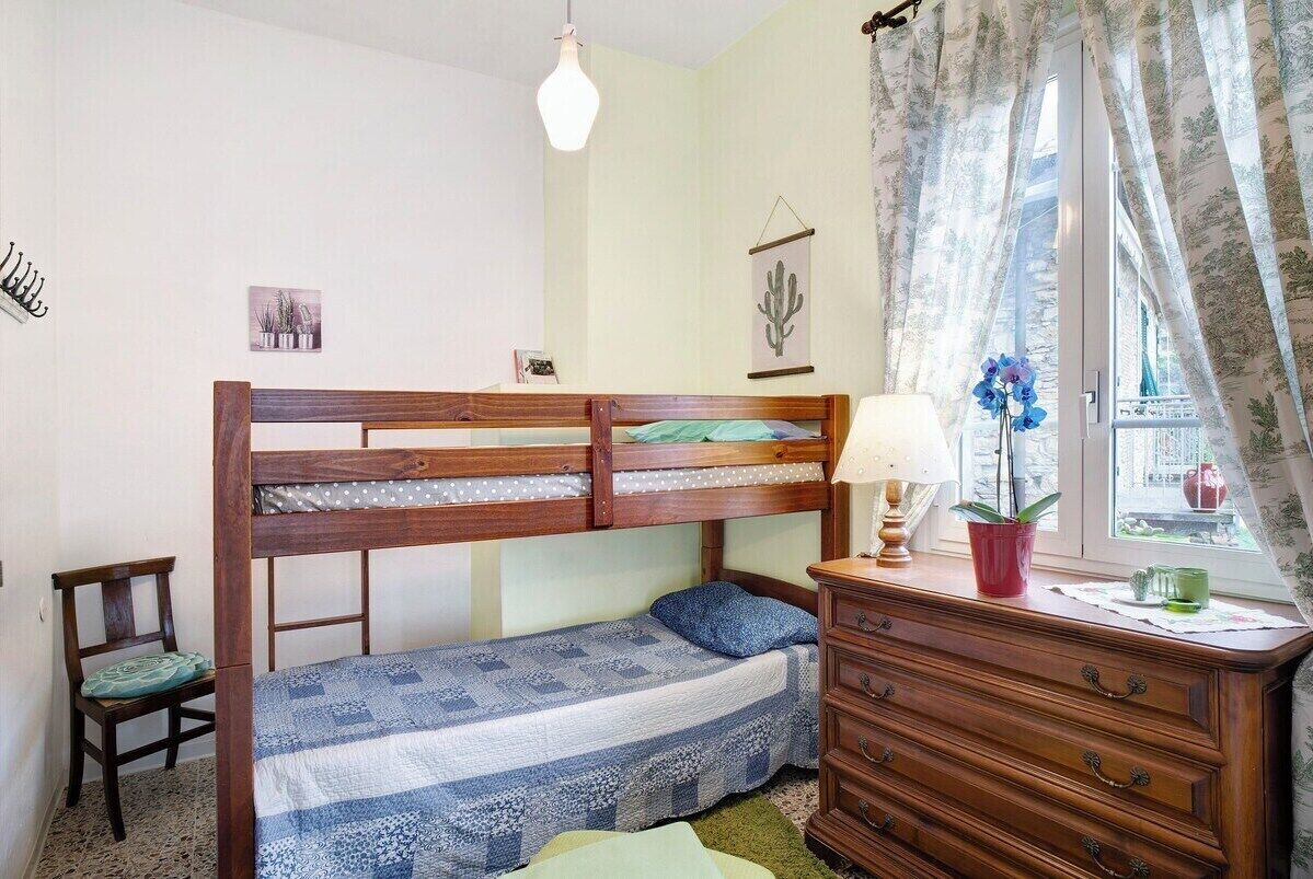 Una cama litera con estructura de madera, una cama individual con colcha azul y blanca, un aparador de madera con cajones, una lámpara y una ventana con cortinas.