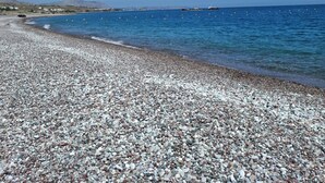 Een kiezelstrand met helderblauw water en land in de verte.