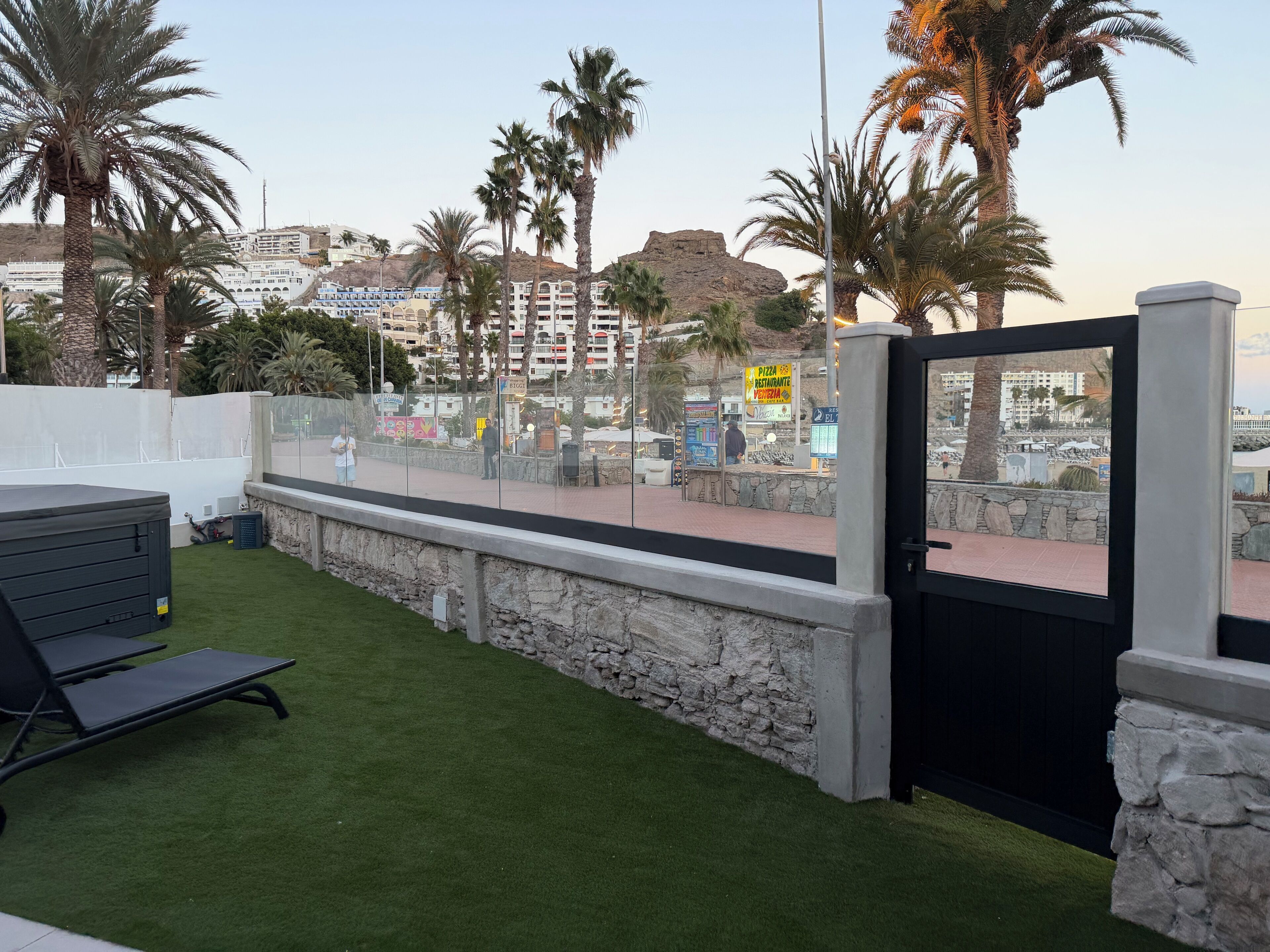A rooftop terrace with artificial grass, a stone wall, and a view of a cityscape with buildings and palm trees.