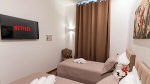 A hotel room with two beds, a TV displaying the Netflix logo, a wall-mounted artwork, and a window with curtains. - Cozy twin room with private bathroom. (Livorno)