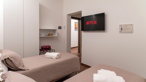 A hotel room with two beds, a TV mounted on the wall, a shelf with items, and a door leading to another room. - Cozy twin room with private bathroom. (Livorno)
