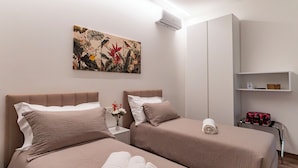 A hotel room with two beds, a wall-mounted air conditioner, a shelf with a basket, and a floral artwork. - Cozy twin room with private bathroom. (Livorno)