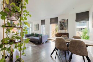 A modern living room with a sofa, dining table, and chairs. There is a large window with a view of the outside. - Sleek 1BDR Flat with Balcony Private Work Space (Greater London)