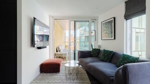 A modern living room with a grey sofa, a red ottoman, a small white table, and a large window with a view of buildings. - Sleek 1BDR Flat with Balcony Private Work Space (Greater London)