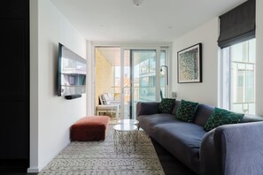 A modern living room with a grey sofa, a red ottoman, a small white table, and a large window with a view of buildings. - Sleek 1BDR Flat with Balcony Private Work Space (Greater London)