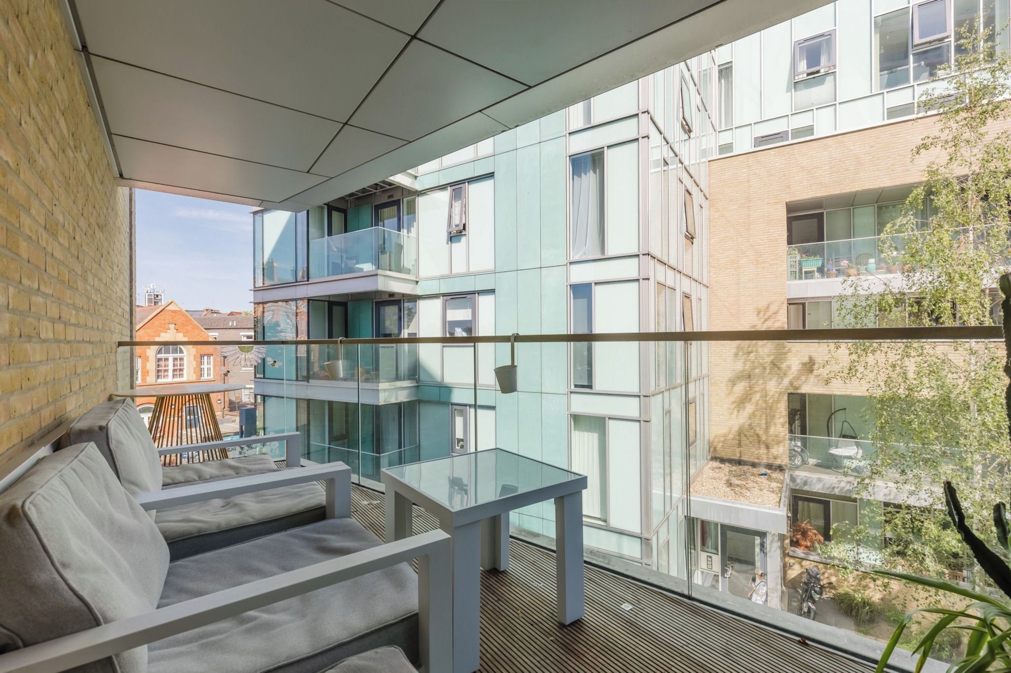 Ein moderner Balkon mit Glasgeländern, einer Sitzgelegenheit mit Kissen und Blick auf angrenzende Gebäude.