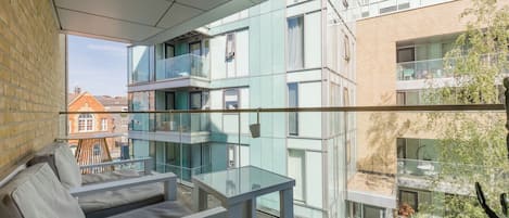 Un balcón moderno con barandillas de vidrio, una zona de estar con cojines y vistas a edificios cercanos.
