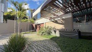 A modern building with a covered outdoor area, a sign reading 'GAMEIRA,' and a paved pathway with greenery.