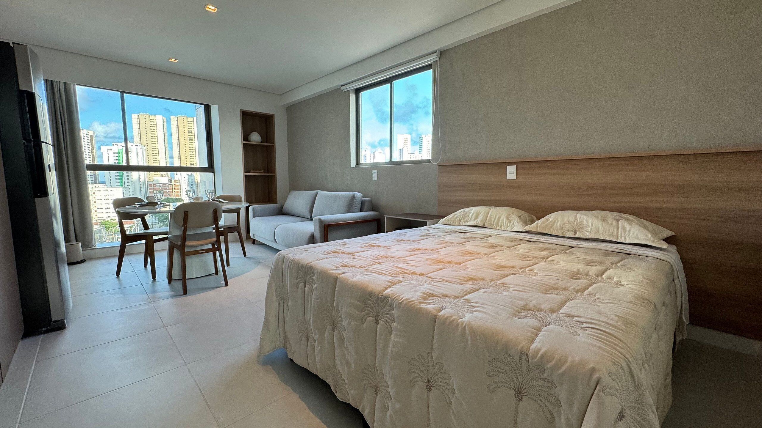 A hotel room with a bed, a desk, a chair, a sofa, and large windows offering a city view.