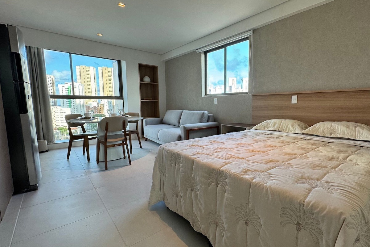 Quarto de hotel com cama, escrivaninha, poltrona, sofá e amplas janelas com vista para a cidade.