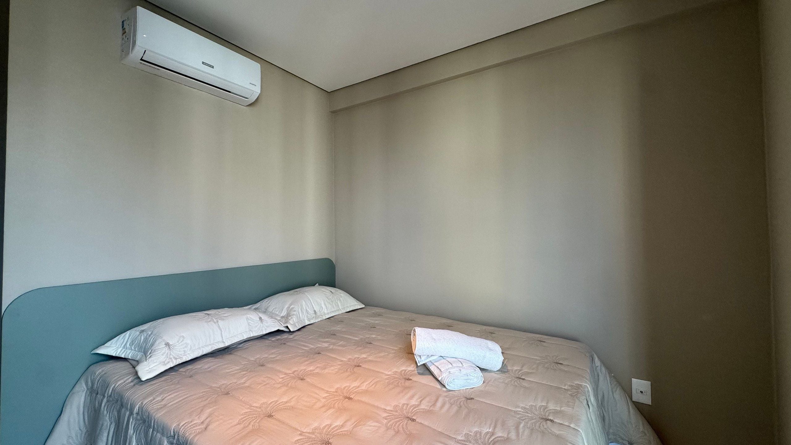 Uma cama de solteiro com cabeceira, uma toalha enrolada e um aparelho de ar condicionado fixado na parede.