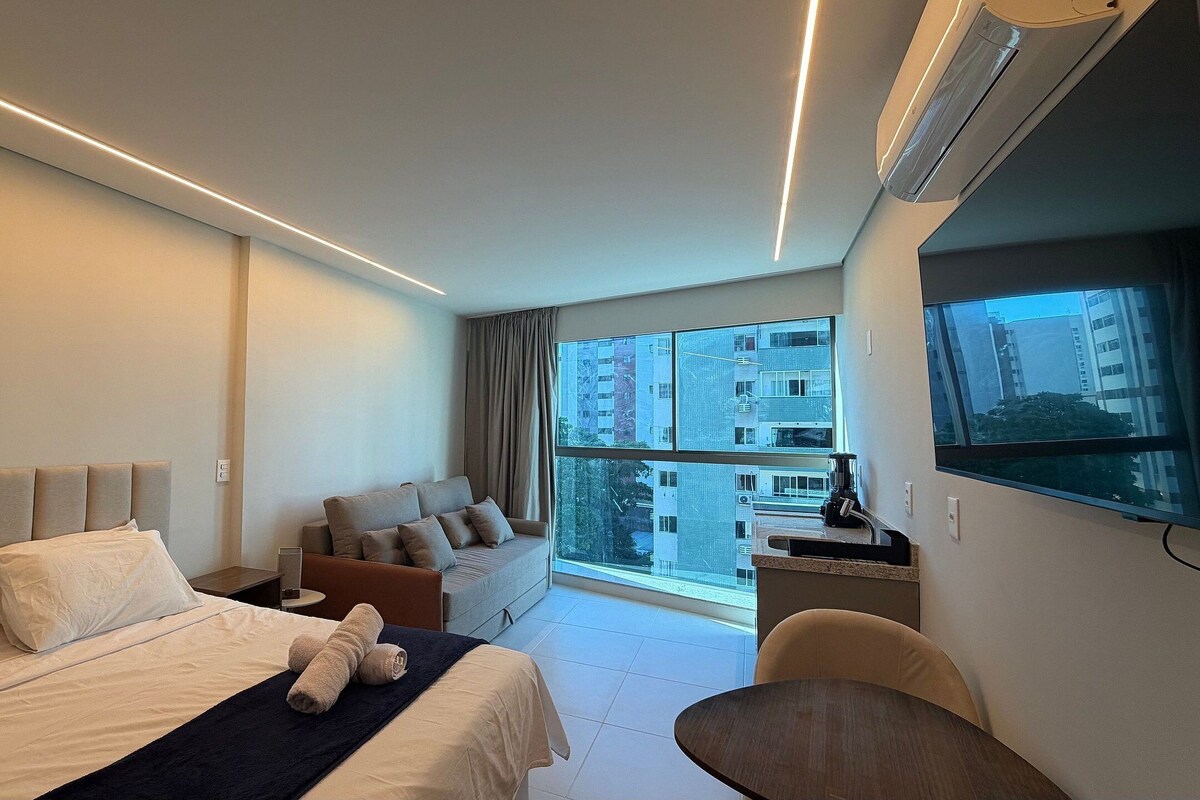 A modern hotel room with a large bed, a sofa, a TV, and a view of buildings.
