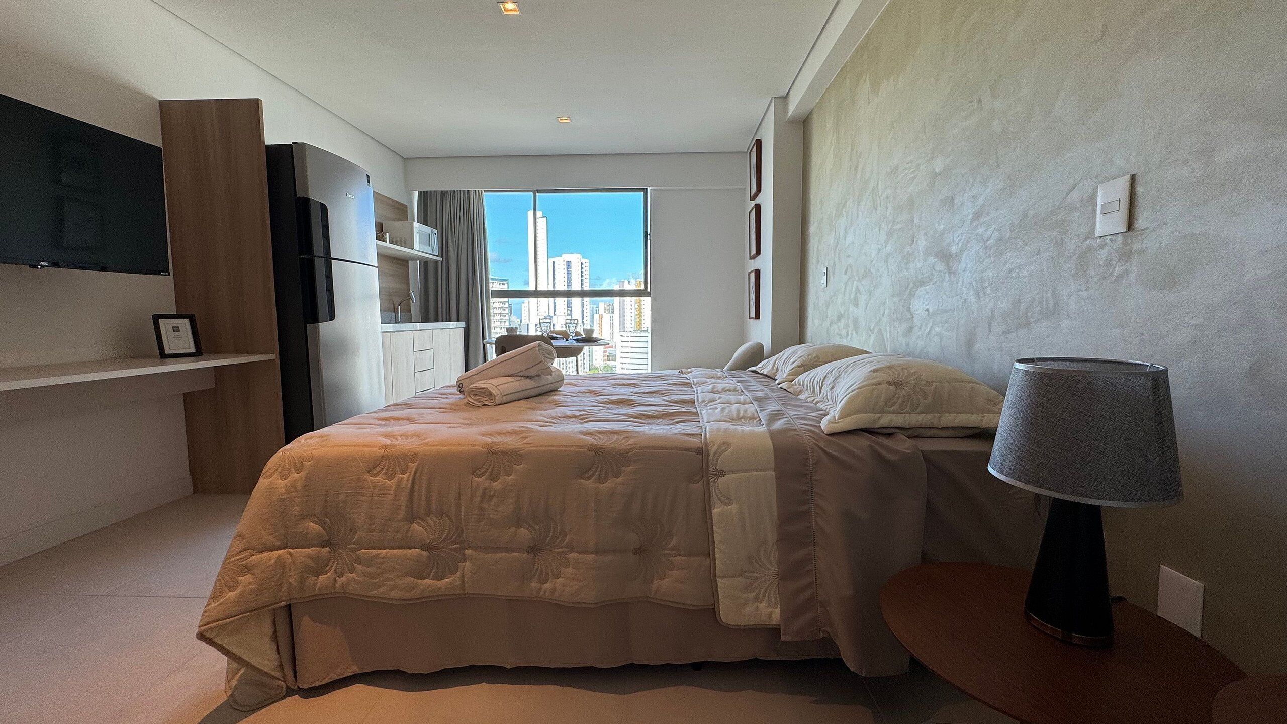 A bedroom with a bed, a lamp, a TV, a window with a city view, and a kitchenette.