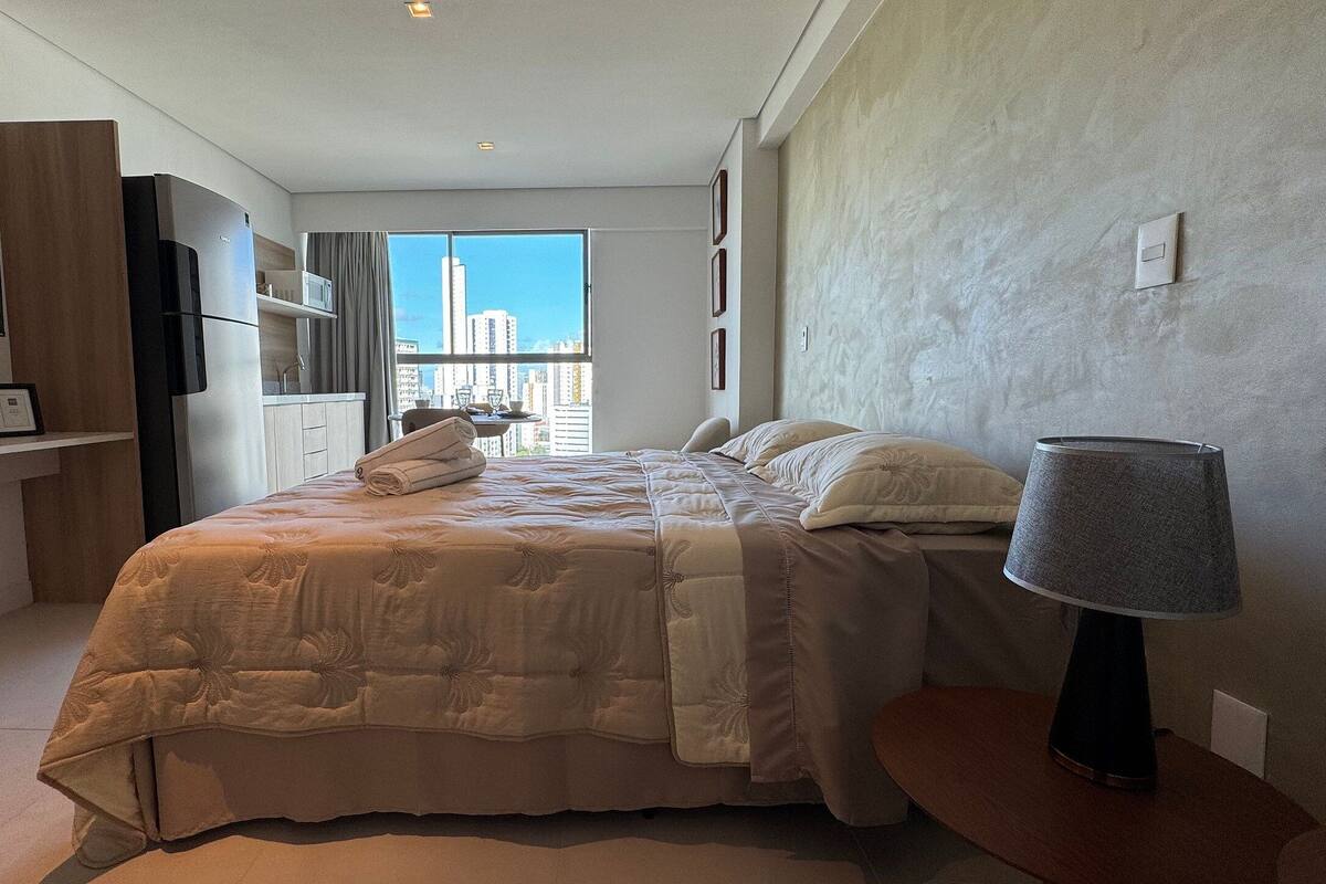 Quarto com cama, luminária, televisão, janela com vista para a cidade e cozinha compacta.