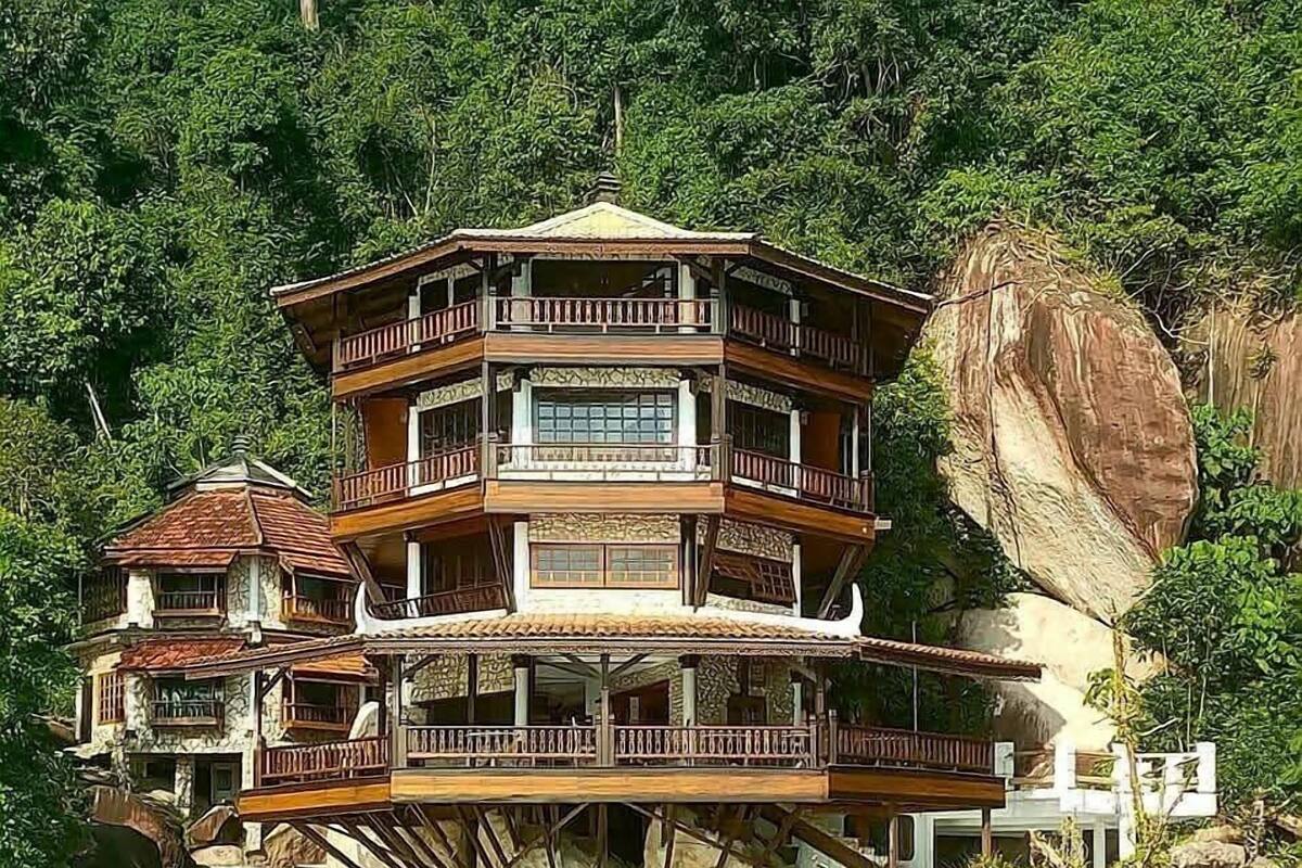 A multi-story building with balconies, built on stilts over water, surrounded by greenery.