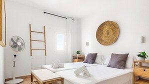 Hotelkamer met twee bedden, een ventilator, een houten ladder, een geweven wanddecoratie en een klein tafeltje met een plant.